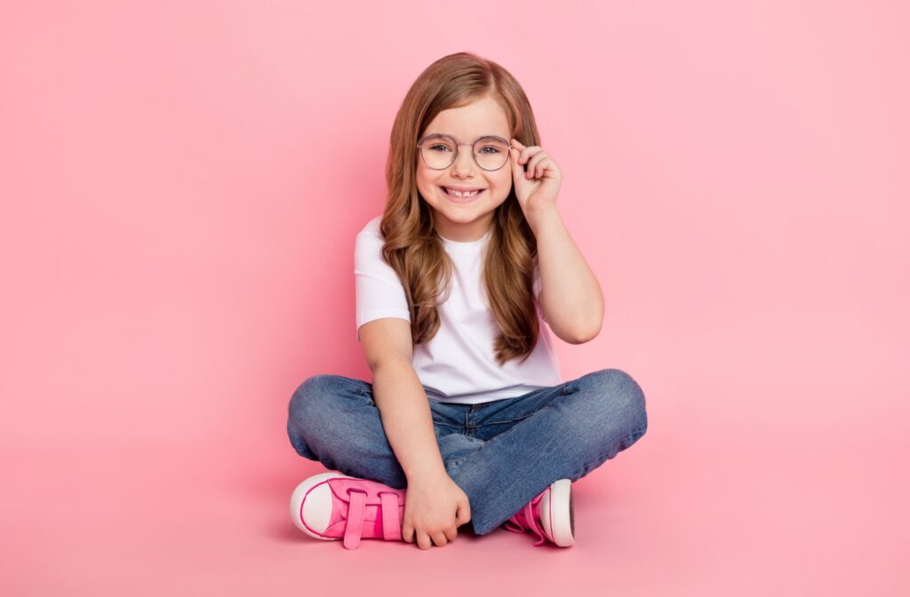 Young smiling girl with glasses sitting cross legged in front of a pink background.