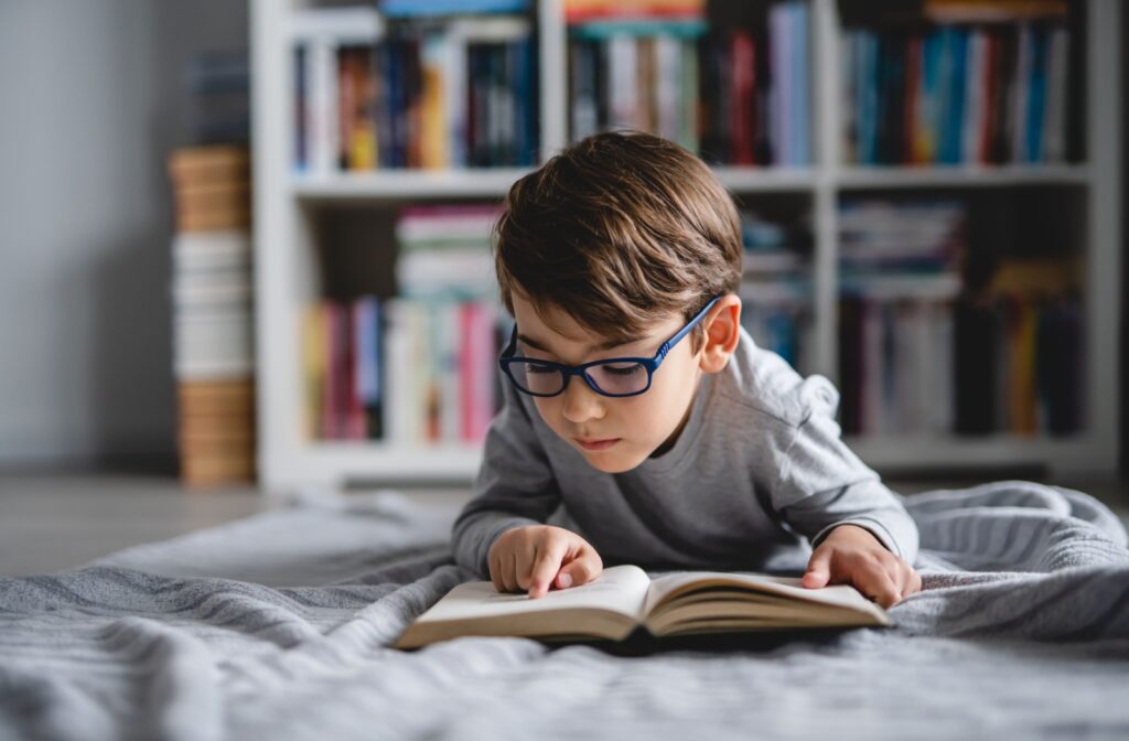 Young boy wearing glasses while lying down and reading a book, supporting eye health and early literacy.