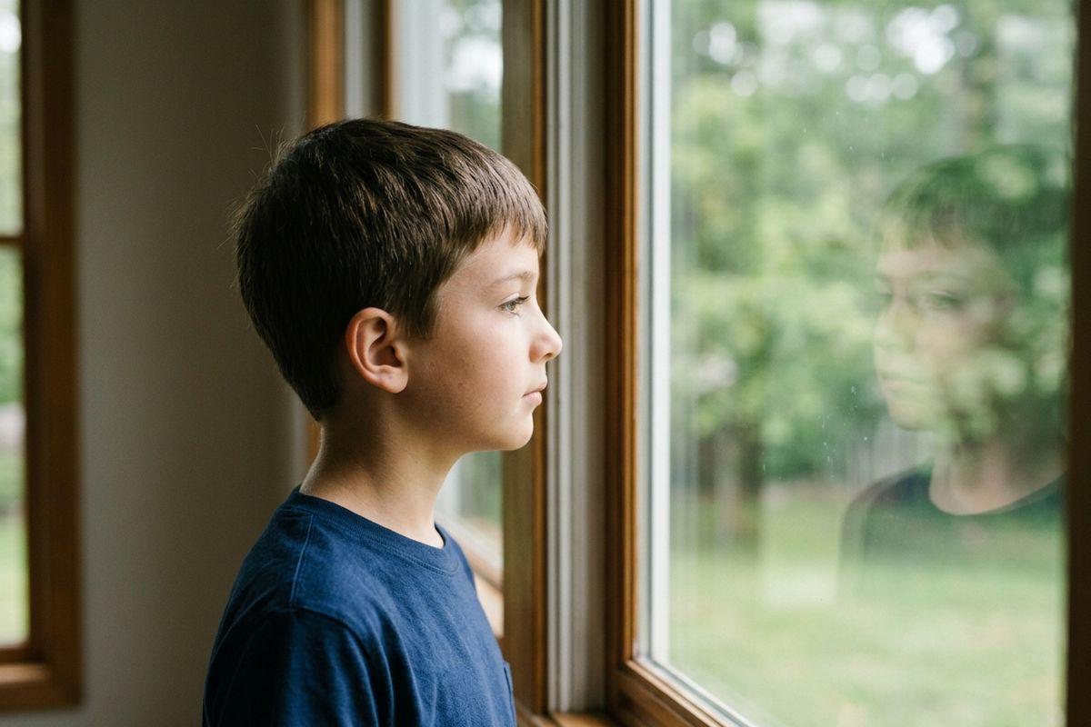 Child in a blue t-shirt gazing out a window indoors, reflection visible in the glass, trees visible outside.